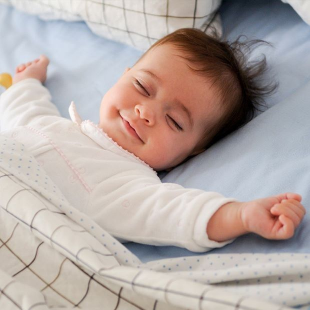 Baby with arms outstretched, eyes closed and smiling lay in a bed